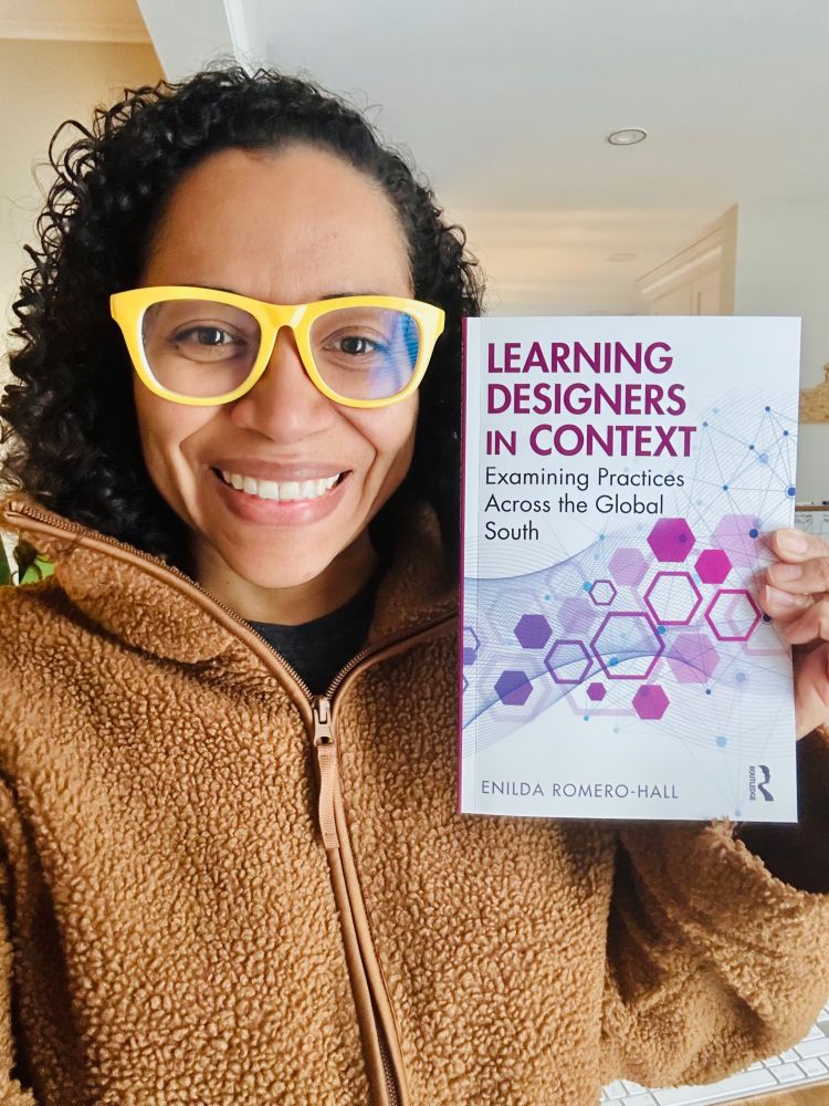A smiling person with curly hair and bright yellow glasses holds up a book titled Learning Designers in Context: Examining Practices Across the Global South by Enilda Romero-Hall. The person is wearing a brown fleece jacket and standing indoors. The book cover features purple and pink hexagonal graphics on a white background.